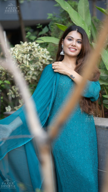 Woman in a teal handmade chikankari suit standing among plants with 'Pakke Dhaage' branding.
