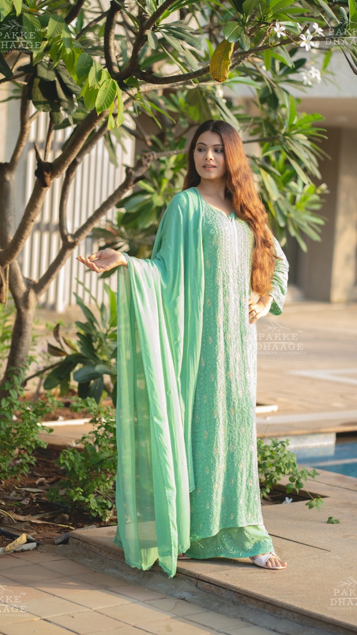 Woman in a light green handmade chikankari outfit standing outdoors near a pool with greenery around.