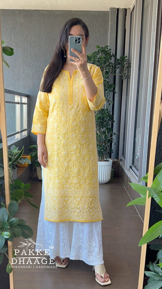 Woman taking a mirror selfie wearing a yellow chikankari kurta with white pants in a room with plants.