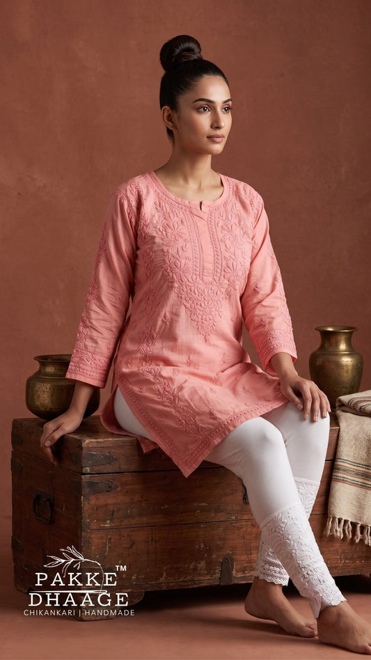 A woman seated on a rustic wooden crate wearing a peach-pink chikankari embroidered kurta with white pants, posed against a warm terracotta backdrop with decorative brass vessels. The brand name Pakke Dhaage Chikankari Handmade is visible in the lower left corner.