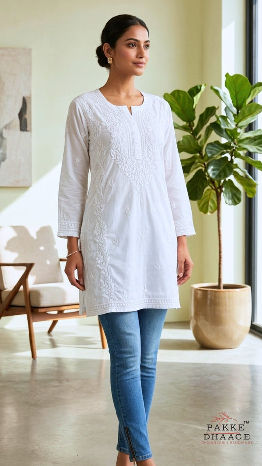Woman wearing a white hand-made chikankari mid length kurta with blue jeans in a bright room with a plant and furniture.