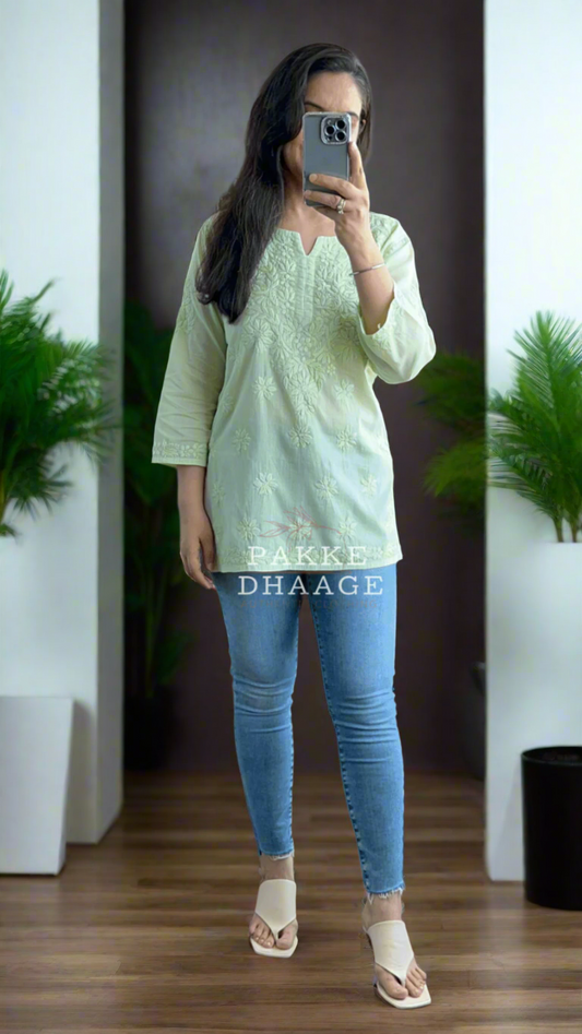 Person wearing a light green chikankari top and blue jeans in a room with plants.
