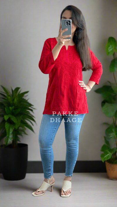 Person wearing a red chikankari top and blue jeans standing in front of a mirror with plants around.