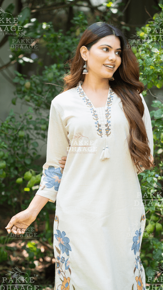 Woman in a cream-colored traditional suit set with floral patterns, standing outdoors with greenery in the background.