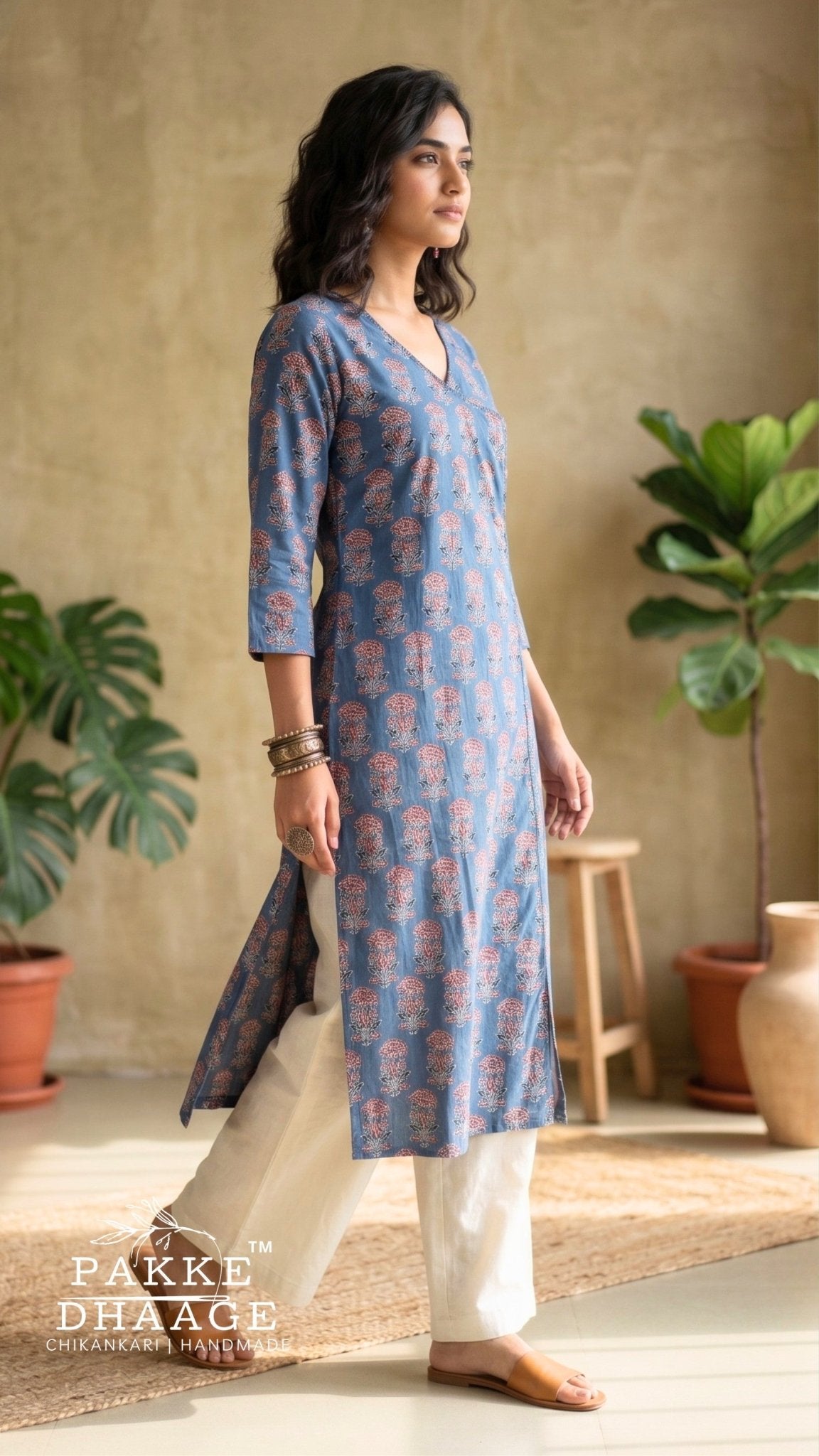 A woman modeling a blue and pink printed kurta with white pants in a rustic indoor setting with potted plants and a wooden stool. The brand logo 'PAKKE DHAGE' is visible in the bottom left corner.