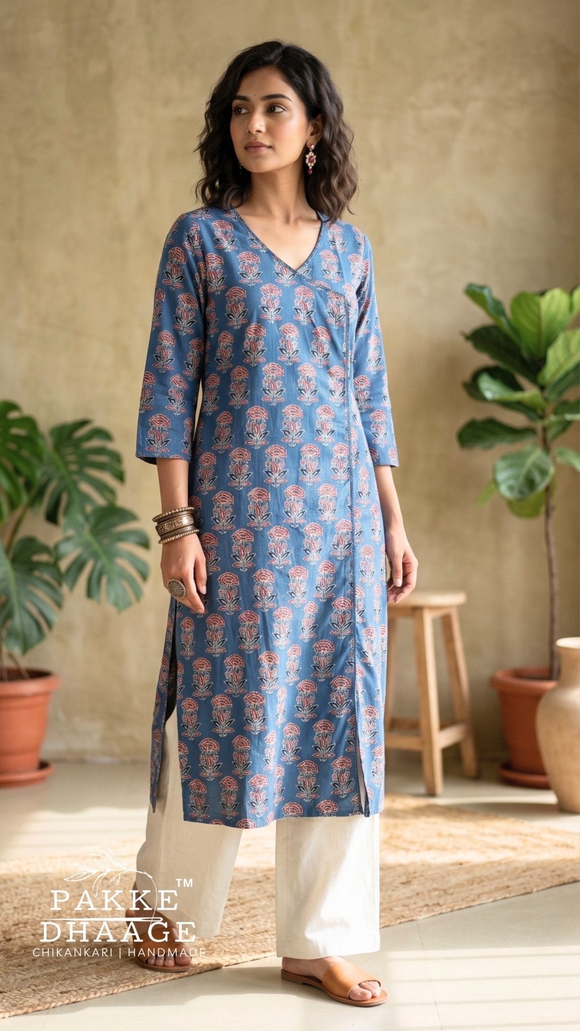 A woman modeling a blue V-neck kurta with a pink and beige floral block print, paired with white wide-leg pants in a warm, rustic indoor setting with potted plants. | Pakke Dhaage