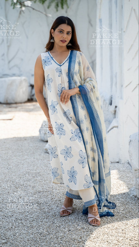 Woman in a floral printed suit in ivory white, with a blue dupatta standing outdoors.
