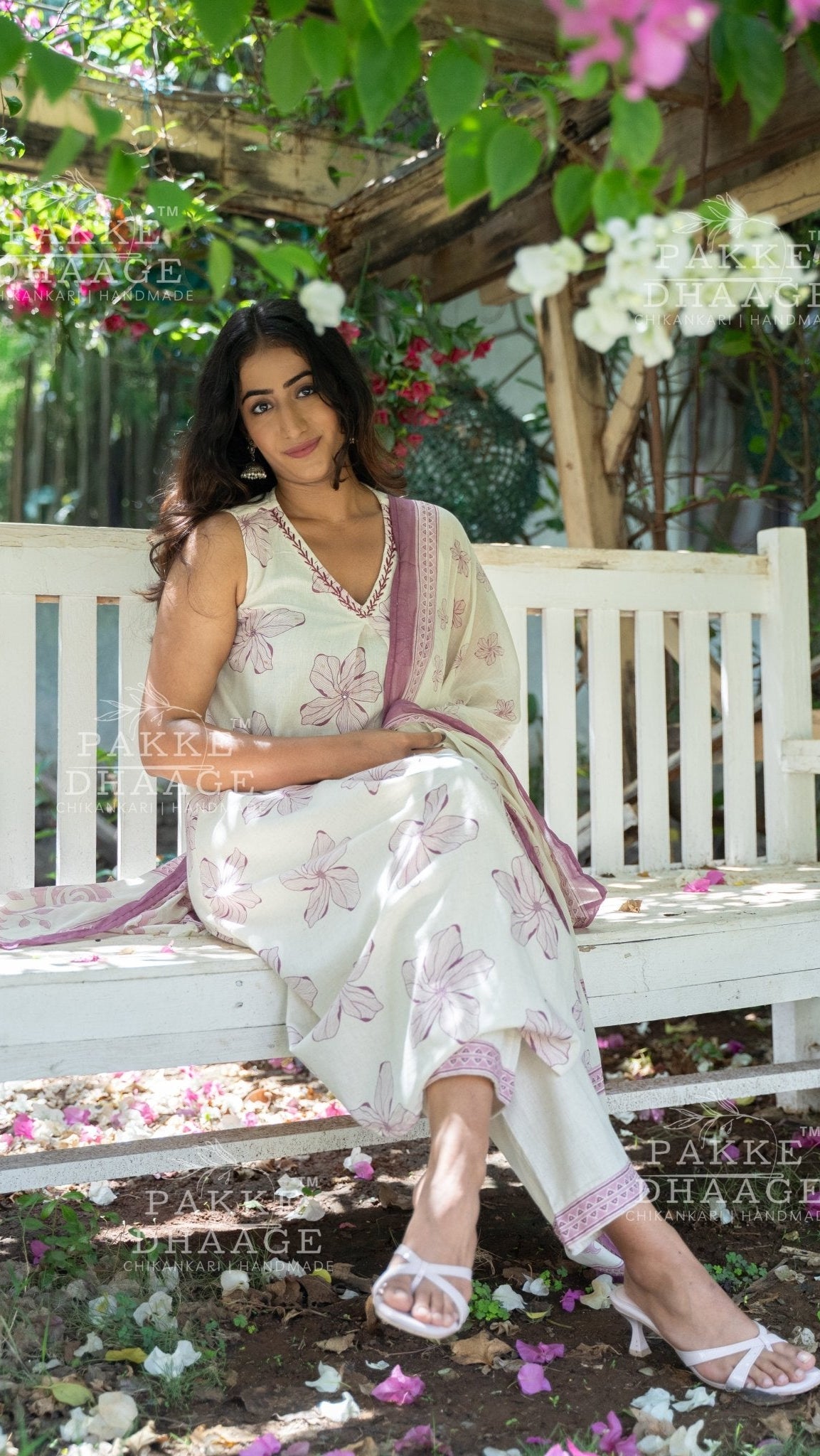 Woman in an ivory white suit set printed with pink floral accents sitting on a white bench in a garden setting.