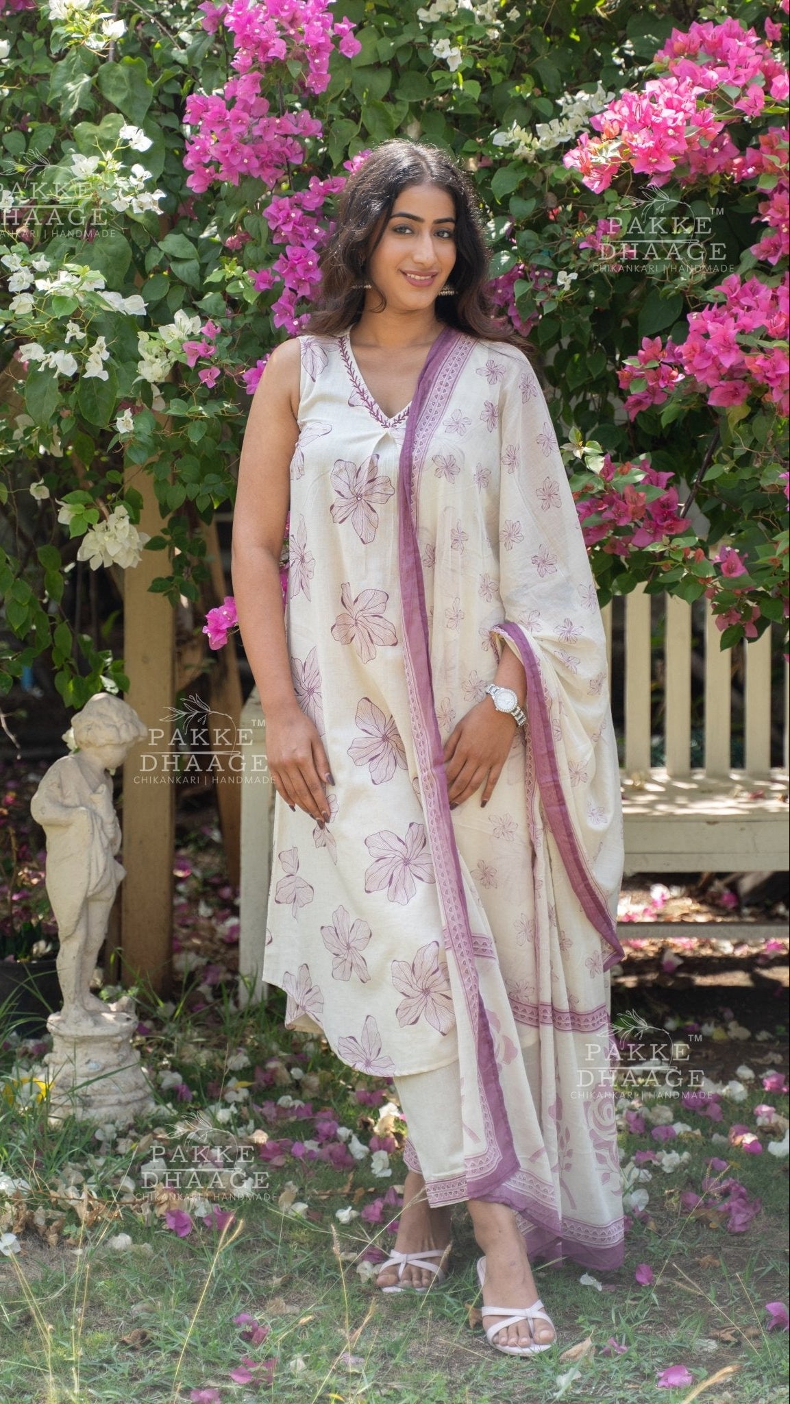 Woman in a white and purple printed suit set, standing in a garden with flowers and a bench.