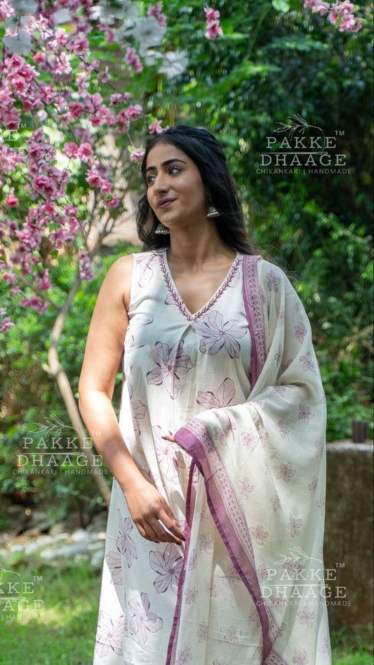 Woman in a white and purple printed & sleeveless suit set standing in a garden with pink flowers.