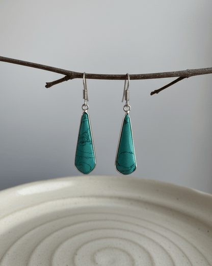Teal drop earrings hanging from a branch on a white plate with a gray background